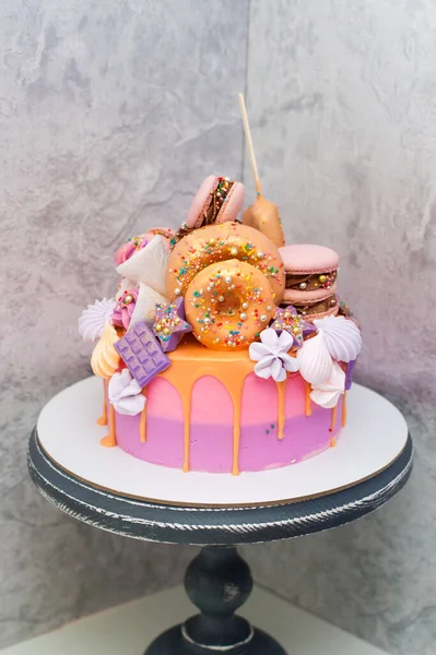 Pink and orange birthday cake decorated with macaroons, meringues, donuts, waffle cones, cake pops and chocolate bars.