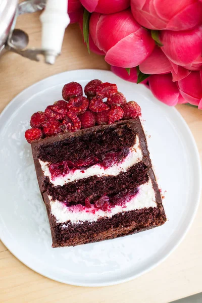 クリームチーズとベリーの充填と新鮮なラズベリーをトッピングしたチョコレートケーキのピース 背景にピンクの牡丹の花束 — ストック写真