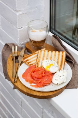 Kızarmış ekmek, tuzlanmış somon ve krem peynirli haşlanmış yumurta ve beyaz pencere kenarında bir fincan latte macciato..