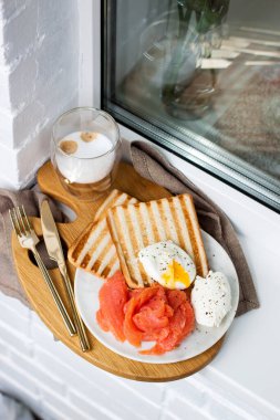 Kızarmış ekmek, tuzlanmış somon ve krem peynirli haşlanmış yumurta ve beyaz pencere kenarında bir fincan latte macciato..