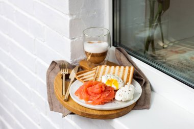 Kızarmış ekmek, tuzlanmış somon ve krem peynirli haşlanmış yumurta ve beyaz pencere kenarında bir fincan latte macciato..
