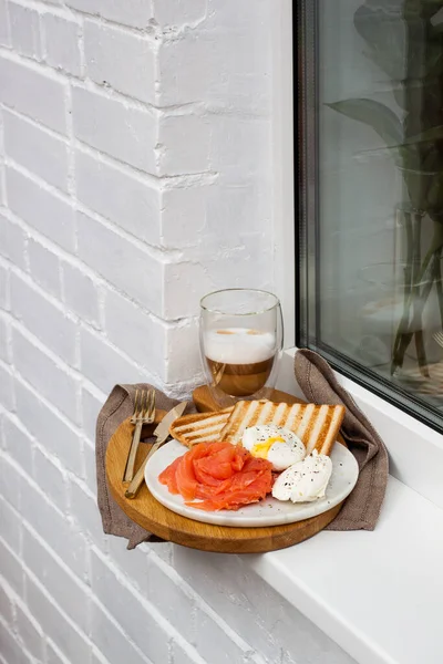 Kızarmış ekmek, tuzlanmış somon ve krem peynirli haşlanmış yumurta ve beyaz pencere kenarında bir fincan latte macciato..