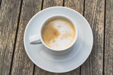 Hot foaming cappuccino in a white ceramich cup on a saucer stands on a wooden table, top view