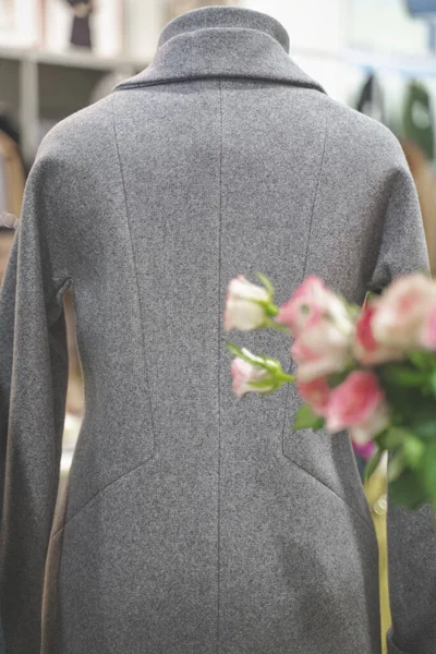 Giysi mağazasındaki bir mankenin üzerine sıcak gri bir ceket giyilir. Ön planda bir buket çiçek var.