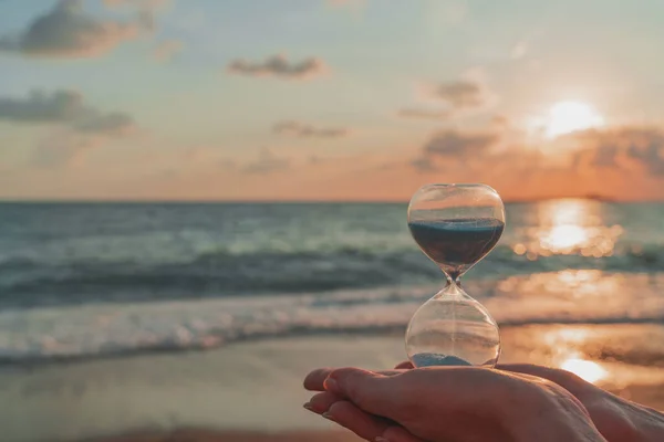 Time is running fast. Hourglass in the hands symbolize the transience of time. Background is seacoast and beautiful red sunset — Stock Photo, Image