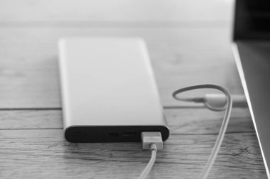 Laptop charging from silver power bank, charger battery on wooden background. Productive portable device for business traveling, black and white image, high angle view and macro