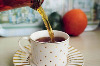 Çaydanlıktan bir fincana aromatik çay dökülüyor, arka planda meyveler var. Fincan ve çay tabağı yerli mutfağın ahşap masasında. Renkli resim ve yakın plan.