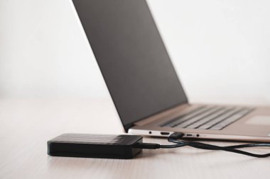 Siyah harici sabit disk, ahşap masanın üzerinde küçük usb kablosuyla dizüstü bilgisayara bağlı, beyaz arka plan. Renkli resim.