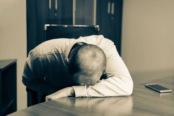 Middle aged man is at the desk in the office, depressed and exhausted. He is just sitting at the working place with his head on the table. Sepia color and horizontal image.