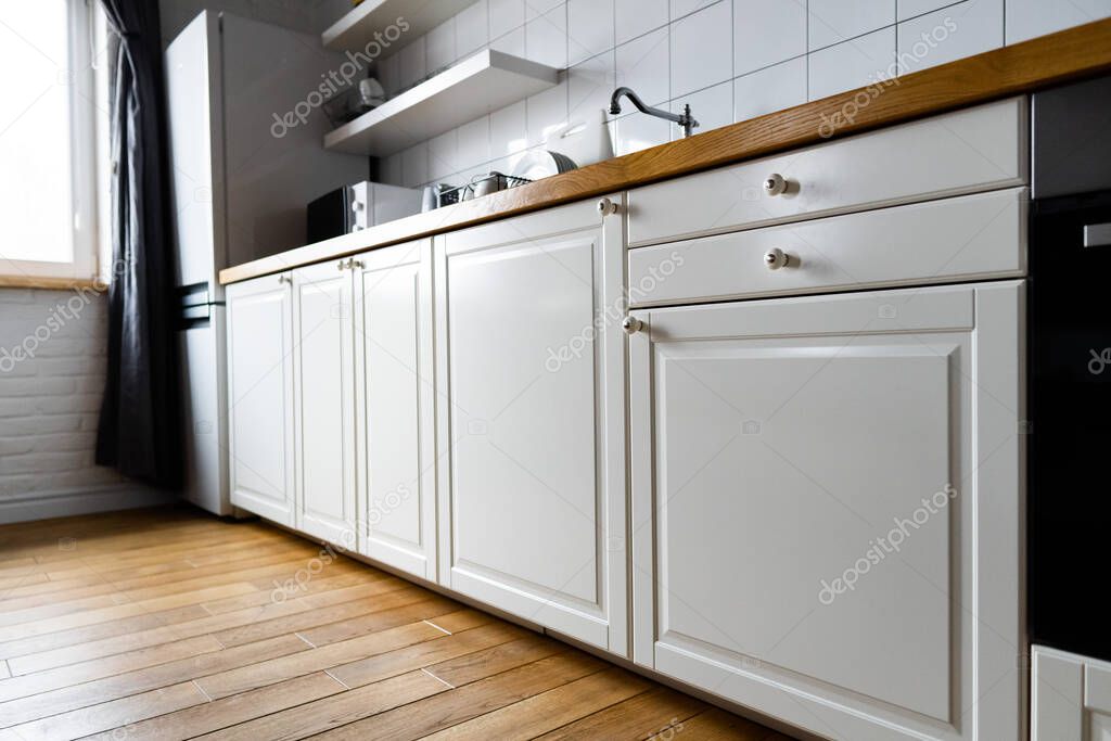Furniture and appliances: bright white cabinets with wooden countertop, electric cooker, induction hob, faucet, sink and dish rack against hardwood floor and light tile in modern scandinavian kitchen