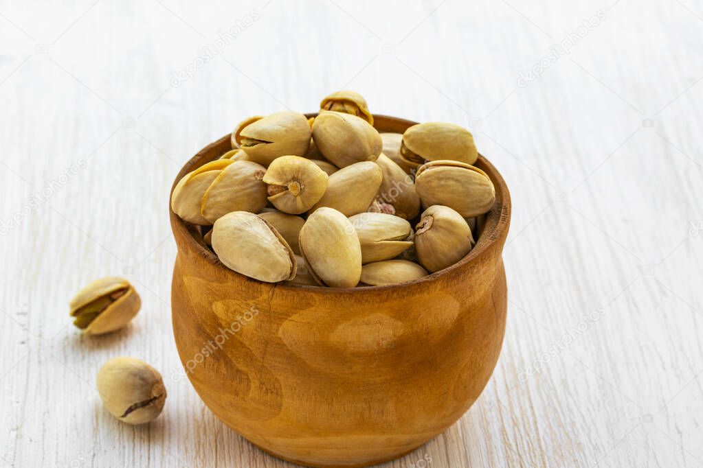 Pistachios lie in a brown wooden bowl on a white wooden background