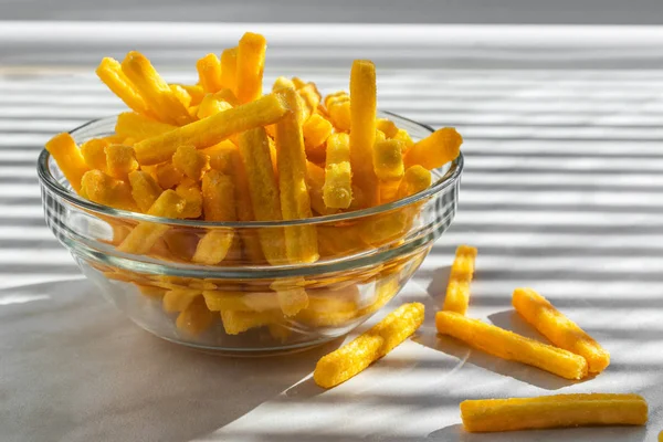 Yellow corn chips in the form of sticks lie in a glass bowl