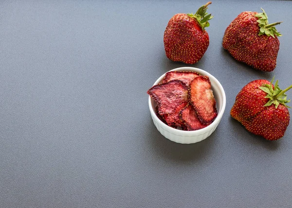 Çilek cipsi beyaz yuvarlak bir fincanda üç çileğin yanında duruyor.