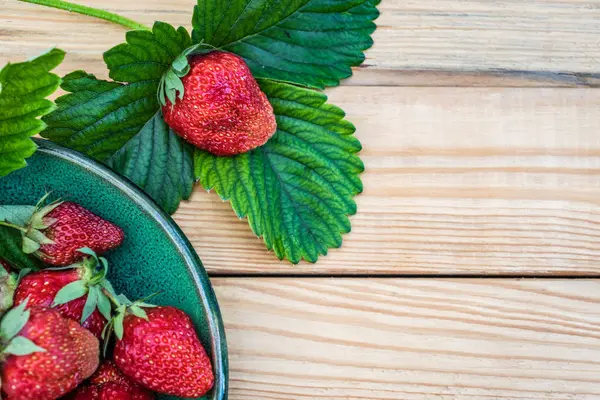 Çilek yemişleri yeşil yaprakların üzerine uzanır. Çileklerin yanında yeşil bir tabakta, tahta tahtaların üzerinde.