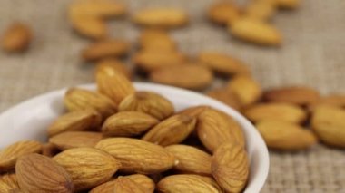 Badem fındıkları beyaz bir kâsede ve gri bir hasır yüzeyde yatar.