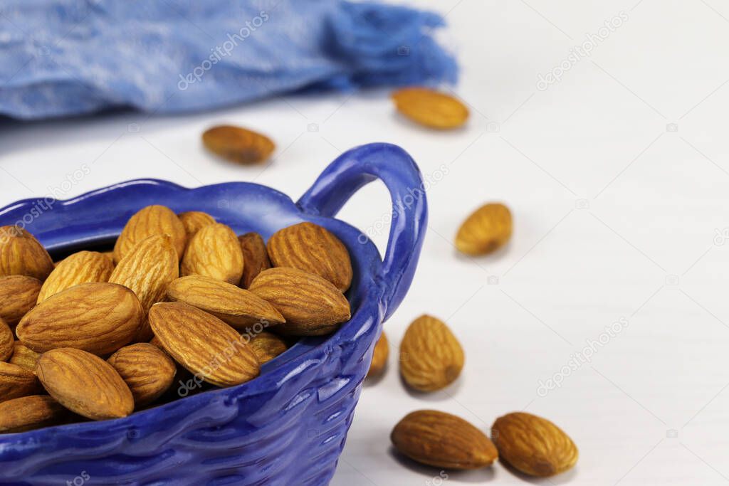 Almond nuts lie in a blue glass basket and on a white surface, close-up