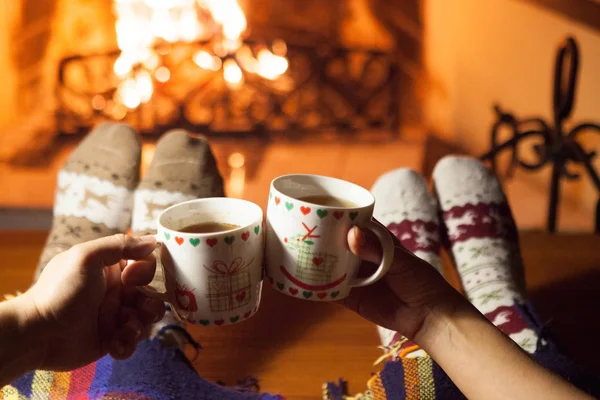 Man and woman in warm knitted socks with cups of hot punch in front of the fireplace