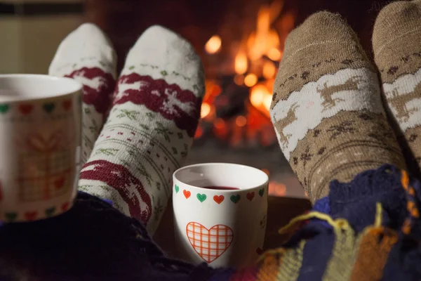 Man and woman in warm knitted socks with cups of hot punch in front of the fireplace