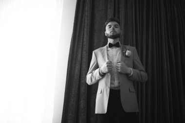 Ceketli şık damat pencerede duruyor. Damat harcı. Düğün fotoğrafçılığı.