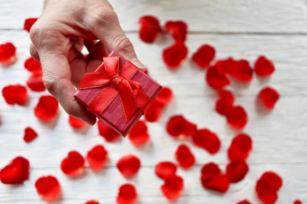 Male hand with a gift box on a background of red rose petals. St. Valentine\'s Day background.