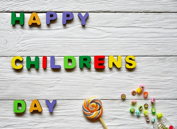 Multi-colored wooden letters making up the words Happy children's day and multi-colored sucking sweets on a white wooden background. Top view. Bright festive background.