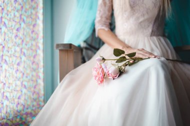 Muhteşem beyaz elbiseli kız elinde çiçeklerle pencerenin kenarında oturuyor.