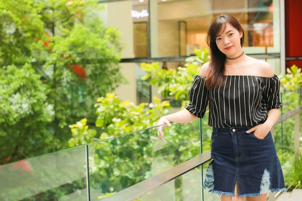 Portrait of Asian beautiful girl wears black off shoulder top and jeans skirt standing by the glass balcony with garden in building. Asian girl model outdoor.