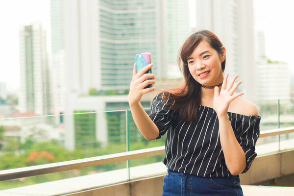 Portrait of happy young girl making video calling and say hi with her friend using mobile phone at rooftop in Bangkok