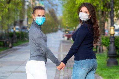 Genç kızlar koruyucu tıbbi maskeler içinde el ele tutuşuyorlar. Kadınlar el ele tutuşur. Eşcinsel ilişkiler kavramı, LGBT insanları, lezbiyenler, salgın, virüs, koronavirüs.