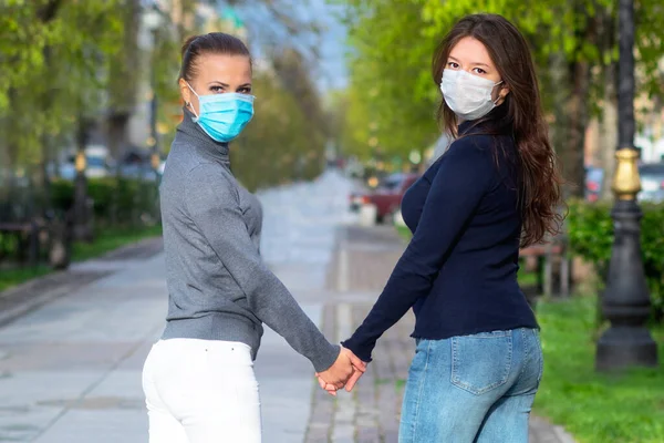 Genç kızlar koruyucu tıbbi maskeler içinde el ele tutuşuyorlar. Kadınlar el ele tutuşur. Eşcinsel ilişkiler kavramı, LGBT insanları, lezbiyenler, salgın, virüs, koronavirüs.