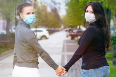 Genç kızlar koruyucu tıbbi maskeler içinde el ele tutuşuyorlar. Kadınlar el ele tutuşur. Eşcinsel ilişkiler kavramı, LGBT insanları, lezbiyenler, salgın, virüs, koronavirüs.