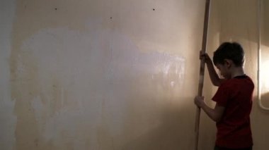 Çocuk uzun kabzalı bir silindirle duvarı beyaz boyayla boyuyor. Video görüntüsü. Arkadan vurulmuş. 4k