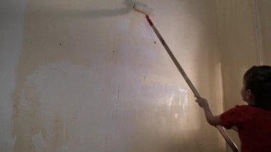 Çocuk uzun kabzalı bir silindirle duvarı beyaz boyayla boyuyor. Video görüntüsü. Arkadan vurulmuş. 4k