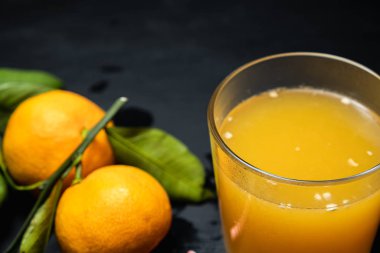 Karanlık bir yüzeyde bir bardak meyve suyunun yanında yaprakları olan üç mandalina vardır..