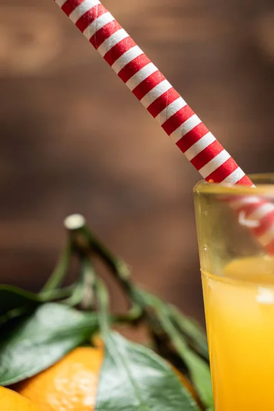 red and white striped straw made not of plastic out in a glass with juice tangerines with leaves lie nearby.