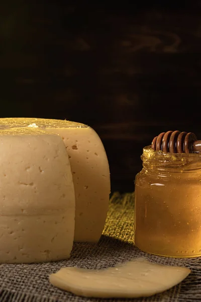 チーズの頭は、ラフラップに横たわるスレート料理にあります。近くには蜂蜜のステンドグラスの瓶があります。ポックマークでスライスされたチーズ。暗い写真 — ストック写真