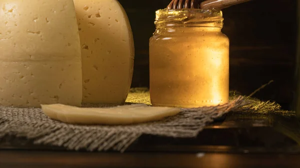 The head of cheese lies on a slate dish lying on a rough burlap. Nearby is a stained jar of honey in which the Honey Stir Bar. A piece of cheese sliced by a pockmark. Low-key lighting.