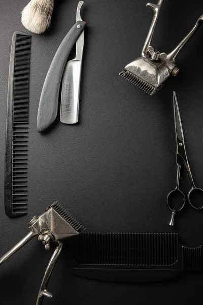 On a black surface are old hairdresser tools. Two vintage hand-held hair clippers, combs, razor, hairdressing scissors, shaving brush. black monochrome. vertical orientation. top view, flat lay