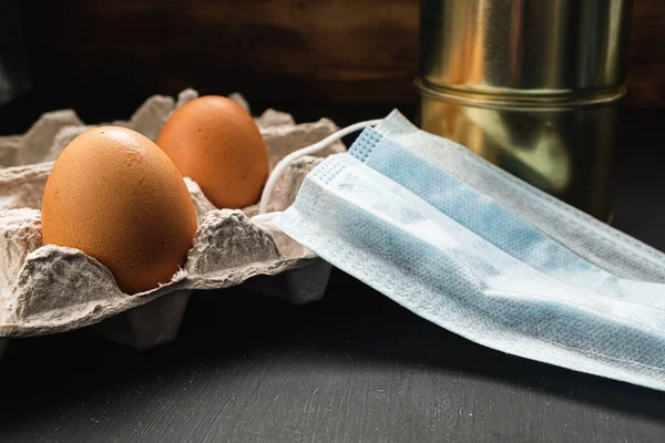 medical mask and two cans of canned food and two eggs in a package on a wooden background.