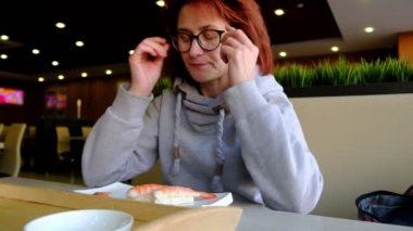 Büyük pencerenin önündeki kafede sushi yiyen kızıl saçlı kız. Açık tenli. Yüzüne gözlük. Kafe 'nin içi. Doğal ışık.