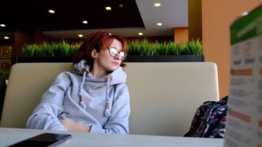 Kafedeki büyük pencerenin önündeki kızıl saçlı kız gözlerini kaçırıyor. Açık tenli. Yüzüne gözlük. Kafe 'nin içi. Doğal ışık.