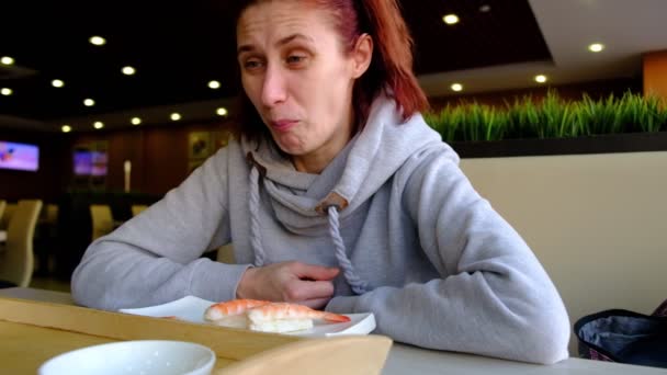 Chica pelirroja en el café en la ventana grande disgustado comer sushi. Piel clara. gafas en la cara. El interior de la cafetería. Luz natural . — Vídeo de stock