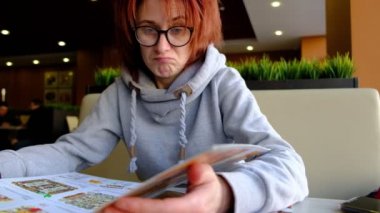 Büyük pencerenin önündeki kafede açık tenli menüye bakan kızıl saçlı kız. Yüzüne gözlük. Kafe 'nin içi. Doğal ışık.