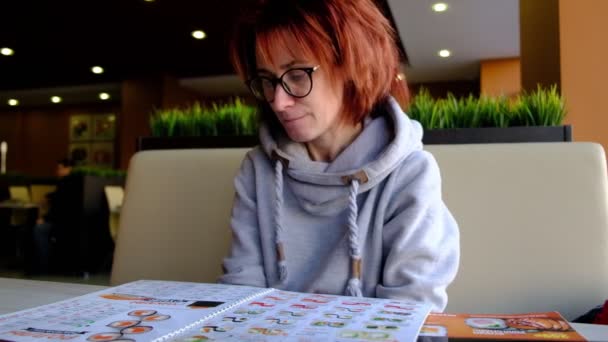 Rothaarige Mädchen im Café am großen Fenster mit Blick auf die Speisekarte Helle Haut. Brille auf dem Gesicht. Das Innere des Cafés. Natürliches Licht. — Stockvideo