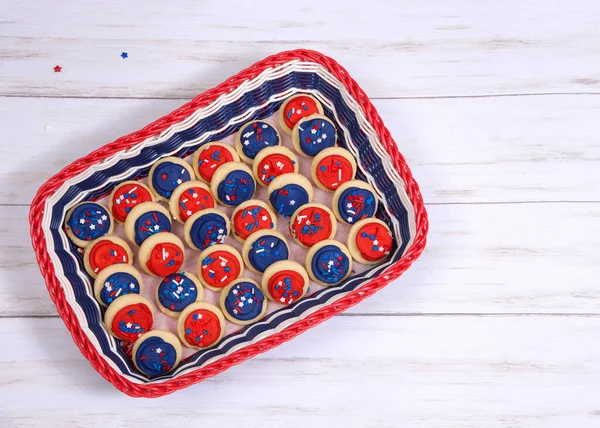 Patriotic basket in red, white and blue filled with bite sized sugar cookies frosted in red and blue frosting covered with stars and sprinkles candies, light wood table with copy space.