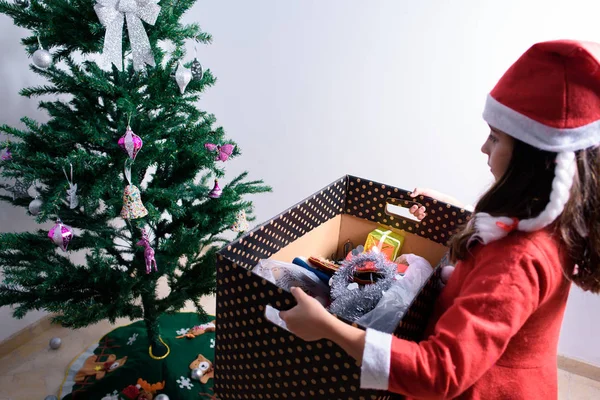 Liten flicka flyttar låda med juldekorationer för att sätta dem på trädet. Klädd som jultomten — Stockfoto