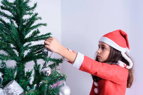 En liten flicka som dekorerar julgranen. Förklädd till jultomten. — Stockfoto