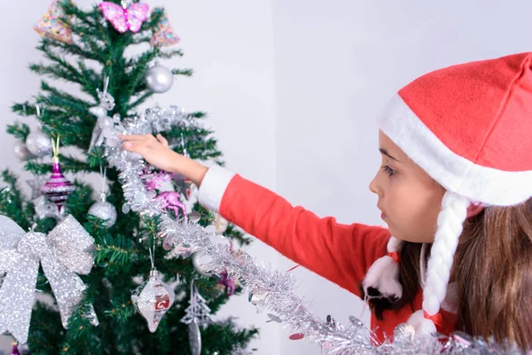 En liten flicka som dekorerar julgranen. Förklädd till jultomten. — Stockfoto