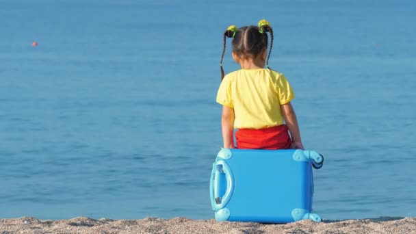 Liten söt flicka i färgglada kläder sitter på en blå resväska på havet stranden. — Stockvideo