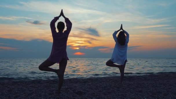 Dos mujeres jóvenes practican yoga junto al mar o el océano al atardecer . — Vídeos de Stock